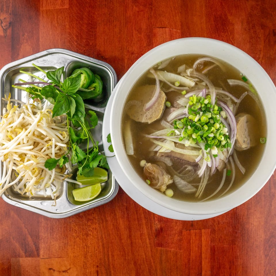 A bowl of pho with meat, noodles, and onions, alongside a tray of bean sprouts, basil, and lime.