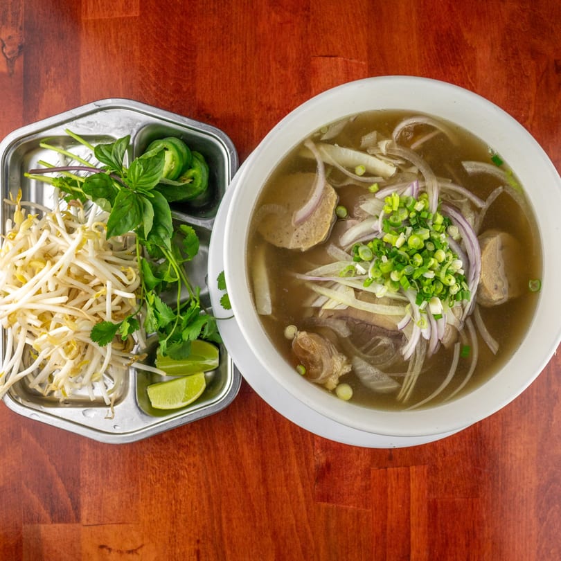 A bowl of pho with meat, noodles, and onions, alongside a tray of bean sprouts, basil, and lime.