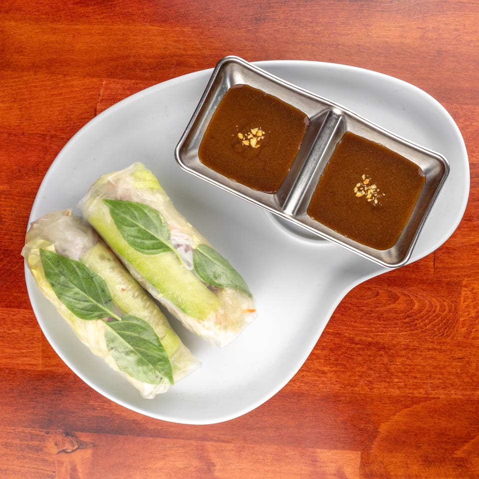 Two fresh spring rolls with basil leaves and a side of peanut dipping sauce on a white plate.