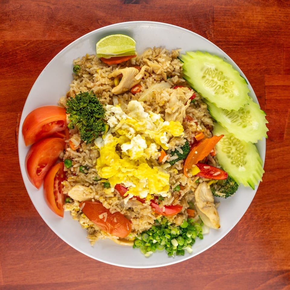 Chicken fried rice with scrambled egg, broccoli, tomatoes, cucumbers, and lime on a white plate.