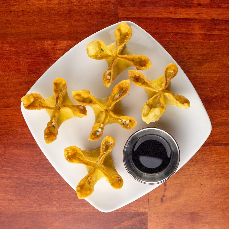 Five golden-fried crab rangoons and a small cup of dark dipping sauce on a white plate.